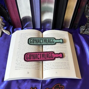 May include: Two rectangular bookmarks with the text "GOYKACTMLAGG" in black, one light blue and one pink, are placed on an open book. The bookmarks are shaped like a bottle. A "Demon Sprinkles" sticker is at the bottom.