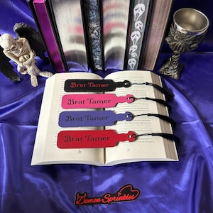 Könnte beinhalten: Vier Leder-Lesezeichen mit dem Text "Brat Tamer" in verschiedenen Farben: Schwarz, Pink, Lila und Rot. Die Lesezeichen befinden sich auf einem Buch mit einem schwarzen Quasten.