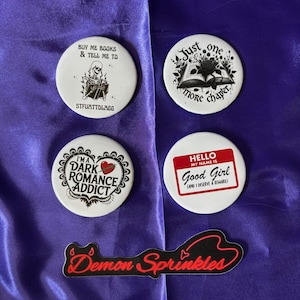 Puede incluir: Cuatro botones blancos y una pegatina negra sobre un fondo morado. Los botones tienen texto y gráficos, incluyendo un esqueleto, un libro abierto y las palabras "Dark Romance Addict". La pegatina dice "Demon Sprinkles".