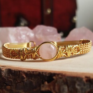 Può includere: Un braccialetto dorato avvolto in filo metallico con una pietra di quarzo rosa. Il braccialetto presenta intricati motivi di filo intrecciato e decorativo. Il quarzo rosa è di colore rosa tenue ed è il punto focale. Il braccialetto è esposto su una superficie di legno.