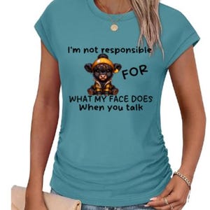 Può includere: T-shirt turchese a maniche corte con la scritta "I'm not responsible FOR WHAT MY FACE DOES When you talk." La maglietta presenta un'illustrazione a cartoni animati di una mucca nera con cappello e giacca gialli.
