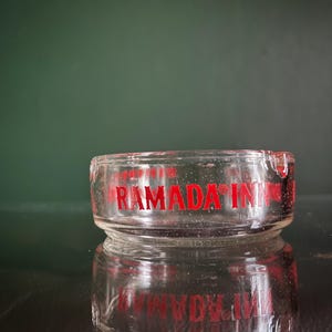 May include: A clear glass ashtray with the words "RAMADA INN" in red. The ashtray is round with a small lip for cigarettes. It sits on a reflective surface. The image is well-lit.