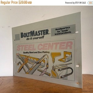 May include: A white metal sign with the text "Boltmaster do-it-yourself" and "Steel Center Quality Steel and Zinc Plating". The sign features a graphic of various metal pieces and tools.