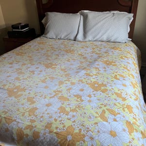 Pode incluir: Uma colcha amarela com um padr&atilde;o floral com flores brancas e laranja. A colcha est&aacute; em uma cama com dois travesseiros brancos.