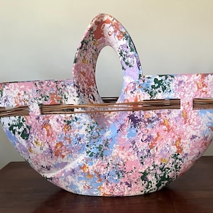 May include: A large, hand-painted basket with a curved handle. The basket is decorated with a colorful splatter pattern in shades of pink, purple, blue, and green. Woven wooden accents are attached to the sides.