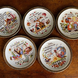 Pode incluir: Conjunto de seis pratos decorativos com borda dourada e desenhos bordados coloridos. Cada prato apresenta um ditado diferente sobre o lar, a cozinha e a felicidade. Os ditados incluem "O lar é onde está o coração", "Beijos não duram, cozinhar sim", "Primeiro o brinde, depois o assado", "Não importa onde eu sirva meus convidados, parece que eles gostam da minha cozinha", "Hoje é o amanhã que você estava preocupado ontem", e "O lar é onde está o coração".