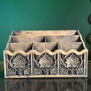 Peut inclure: Un organiseur de bureau en bois avec un motif floral sculpté. L'organisateur comporte trois compartiments et un panneau avant décoratif avec des fleurs et des feuilles sculptées. Le bois a une finition naturelle et vieillie, et l'organisateur repose sur une surface réfléchissante.