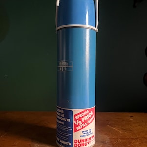 May include: A vintage blue Thermos brand insulated bottle with a white handle. The bottle has a blue cap and a white band. A Dunkin' Donuts label is on the bottom, with text that reads "Special 1/2 Price Refill Offer."
