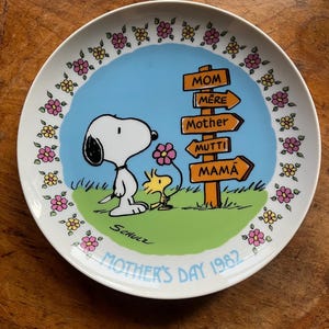 Può includere: Un piatto decorativo con Snoopy e Woodstock con un cartello stradale che indica "Mom", "Mère", "Mother" e "Mutti". Il piatto ha uno sfondo blu con una base verde ed è bordato da fiori rosa e gialli. È presente anche il testo "Mother's Day 1982".