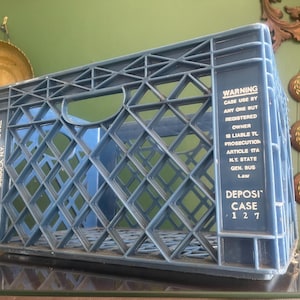 May include: A blue plastic milk crate with a diamond-patterned design. The crate has the text "HONEYWELL FARMS INC. JAMAICA NY" and "WARNING" with legal text. The crate is sitting on a glass surface.