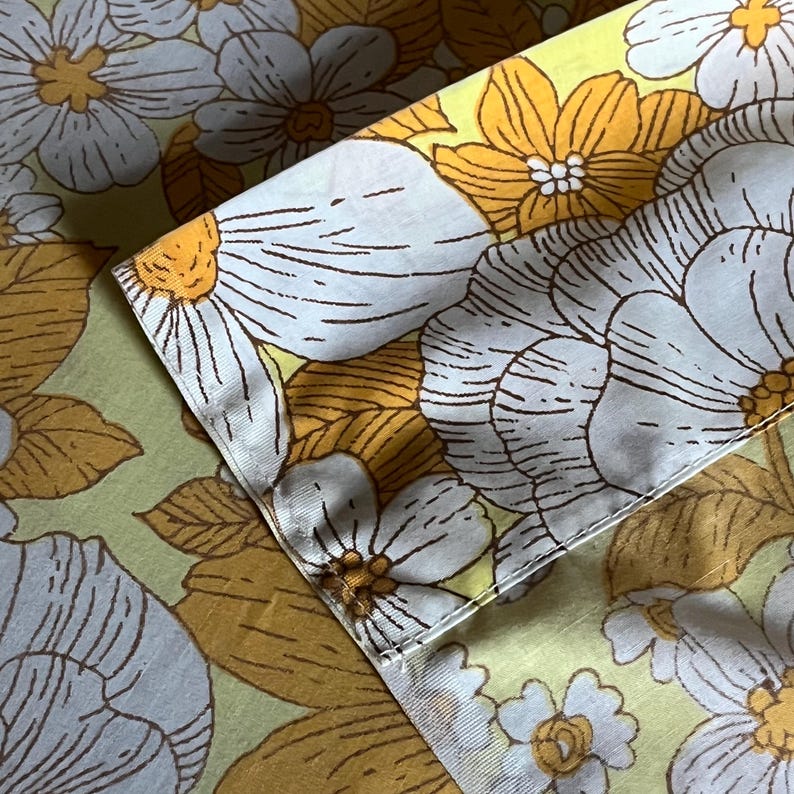Pode incluir: Um close-up de um tecido com um padr&atilde;o floral. O tecido &eacute; de cor verde claro com flores marrons, amarelas e brancas. As flores s&atilde;o estilizadas e t&ecirc;m um aspeto retro.