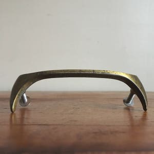 Puede incluir: Un tirador de armario de latón con un diseño curvo en forma de arco. El tirador tiene un acabado cepillado y se fija con tornillos.