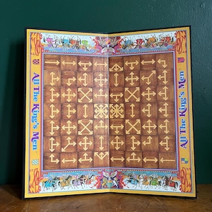 May include: Open board game titled "All The King's Men." The game board is open, revealing a grid of brown squares with directional arrows and symbols. The border is yellow with illustrations of people and horses.
