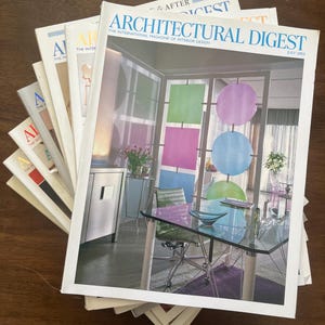 May include: A stack of Architectural Digest magazines. The top magazine features a colorful interior design with a glass table and colorful circles. The magazines are stacked on a dark wooden surface.