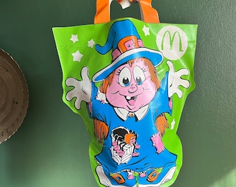 May include: A green and white Halloween-themed treat bag with a witch character holding a spider. The bag has a yellow handle and the McDonald's logo is printed on the front.