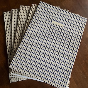 May include: A stack of notebooks with patterned covers. The covers feature a repeating design of the letter "j" in navy blue or brown against a cream background. A blank label is on the front cover of the navy blue notebook.