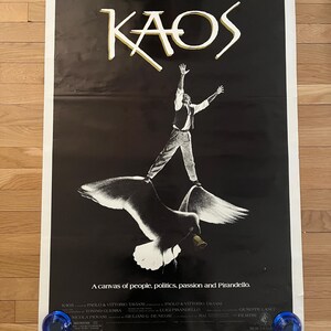May include: A black and white movie poster for the film "Kaos" with the title in gold lettering. The poster features a man standing on a seagull with a bell around its neck. The tagline reads "A canvas of people, politics, passion and Pirandello."