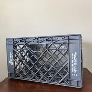 May include: A gray plastic crate with a diamond-patterned design. The crate has the text "Johanna Farms Inc." on one side and "MISUSE OF THIS CASE IS LIABLE TO PROSECUTION" on the other. The crate is sitting on a wooden surface.