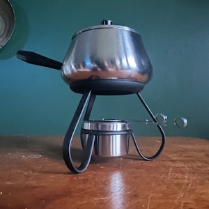 Può includere: Un pentolino per fondue d'argento con manico e supporto neri. Il pentolino è su una superficie di legno.