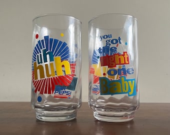 Può includere: Due bicchieri di vetro trasparente Pepsi con grafica colorata. Un bicchiere dice "uh huh" con un design rosso, giallo e blu. L'altro bicchiere dice "you got the right one baby" con un design rosso, giallo, blu e verde.