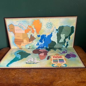 May include: A vintage Risk board game, open to display the world map. The map is colorful, with continents in shades of orange, green, blue, and purple. A ship illustration is in the lower left, and the Risk logo is in the lower right.