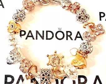 May include: A silver charm bracelet with a variety of charms, including hearts, snowflakes, and a crown. The charms are made of silver, gold, and rose gold. The bracelet is on a white background with the word "PANDORA" printed on it.