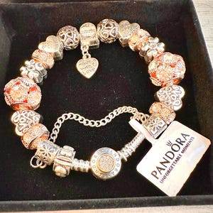 May include: A silver charm bracelet with a variety of charms, including hearts, flowers, and a charm that says "Made with Love". The bracelet is on a black background and has a Pandora tag that says "Unforgettable Moments".