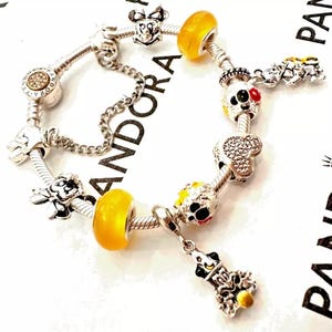 May include: A silver charm bracelet with a variety of Disney-themed charms, including Mickey Mouse, Minnie Mouse, and Goofy. The charms are silver, yellow, red, and black.