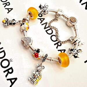 May include: A silver charm bracelet with a variety of Disney-themed charms, including Mickey Mouse, Minnie Mouse, and a yellow glass bead. The bracelet is made by Pandora.