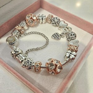 May include: A silver charm bracelet with a variety of charms, including hearts, flowers, and a charm that says "made with love". The bracelet is in a pink box.