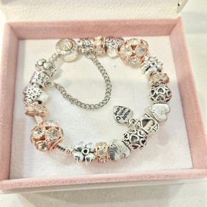 May include: A silver and rose gold charm bracelet with a variety of charms, including hearts, flowers, and a charm that says "Made with love".
