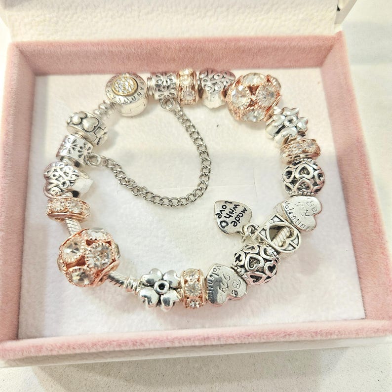 May include: A silver and rose gold charm bracelet with a variety of charms, including hearts, flowers, and a charm that says "Made with love".