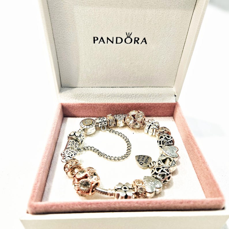 May include: A silver charm bracelet with a variety of charms, including a heart charm that says "Made with Love", a flower charm, and a charm with a small crown. The bracelet is in a white box with the word "Pandora" printed on the lid.