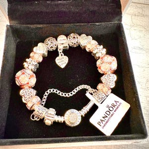 May include: A silver charm bracelet with a variety of charms, including hearts, flowers, and a charm that says "Made with Love". The bracelet is in a black box with a white tag that says "Pandora Unforgettable Moments".