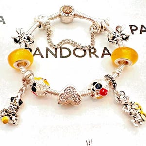 May include: A silver charm bracelet with a variety of charms, including a heart, a Mickey Mouse head, and a yellow glass bead. The bracelet is on a white background with the word "PANDORA" printed on it.