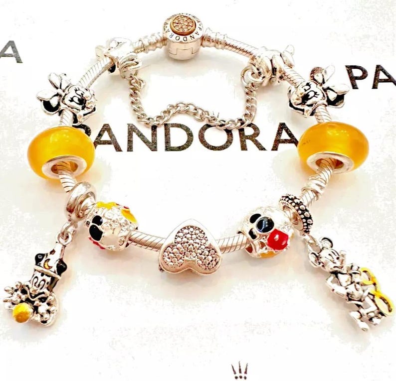 May include: A silver charm bracelet with a variety of charms, including a heart, a Mickey Mouse head, and a yellow glass bead. The bracelet is on a white background with the word "PANDORA" printed on it.