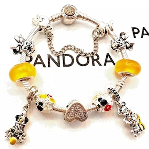 May include: A silver charm bracelet with a variety of charms, including Mickey Mouse and Minnie Mouse characters, a heart charm with clear crystals, and yellow glass beads. The bracelet is on a white background with the word "PANDORA" printed on it.