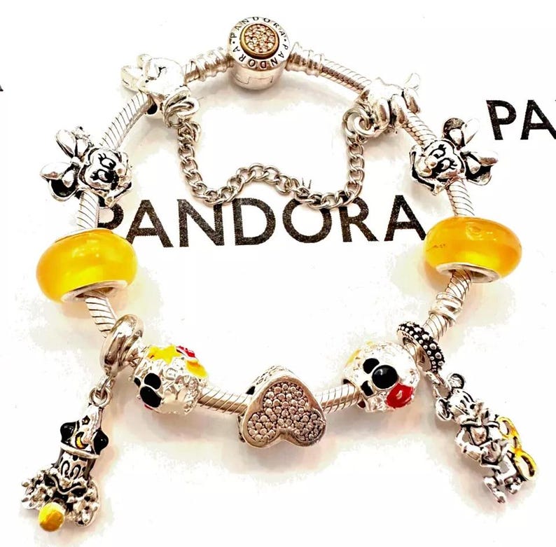 May include: A silver charm bracelet with a variety of charms, including Mickey Mouse and Minnie Mouse characters, a heart charm with clear crystals, and yellow glass beads. The bracelet is on a white background with the word "PANDORA" printed on it.