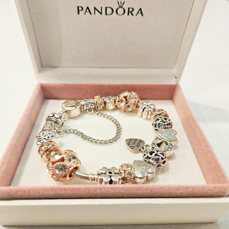 May include: A silver and rose gold charm bracelet with a variety of charms, including hearts, flowers, and a charm that says "Made for you". The bracelet is in a white box with the word "Pandora" on the lid.
