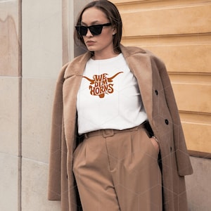 May include: A white t-shirt with a brown graphic of a longhorn bull with the text "We Dem Horns" on it. The person wearing the shirt is standing in front of a brown wooden wall.