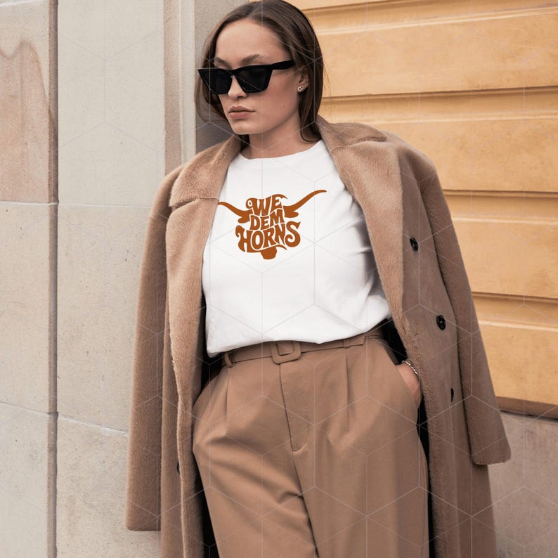 May include: A white t-shirt with a brown graphic of a longhorn bull with the text "We Dem Horns" on it. The person wearing the shirt is standing in front of a brown wooden wall.