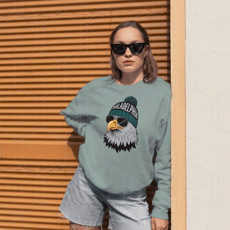 Peut inclure: Un sweat-shirt bleu sarcelle avec un graphique d'un aigle chauve portant des lunettes de soleil et un bonnet avec le mot "PHILADELPHIA" dessus.