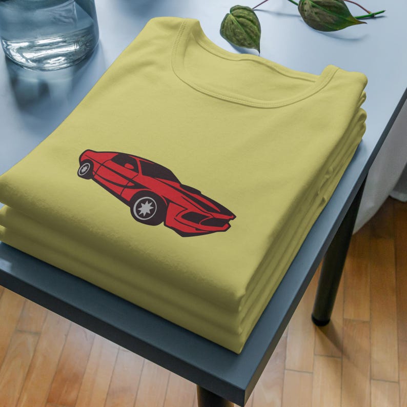Pu&ograve; includere: Una maglietta gialla con una grafica di un'auto d'epoca rossa stampata sul davanti. La maglietta &egrave; piegata e impilata su un tavolo.
