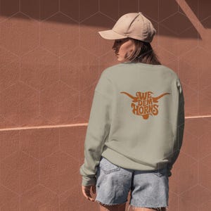 May include: A light green sweatshirt with a brown graphic of a longhorn bull's head with the text "We Dem Horns" on the back. The sweatshirt is being worn by a person wearing a beige baseball cap and denim shorts.