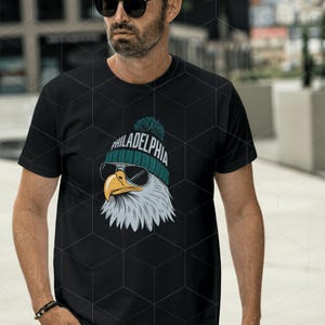 Peut inclure: T-shirt noir avec un graphique d'un aigle chauve portant un bonnet vert et blanc avec le mot "PHILADELPHIA" dessus. L'aigle porte des lunettes de soleil.