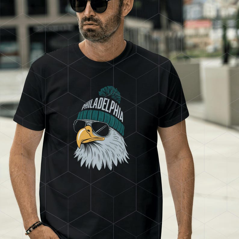 Peut inclure: T-shirt noir avec un graphique d'un aigle chauve portant un bonnet vert et blanc avec le mot "PHILADELPHIA" dessus. L'aigle porte des lunettes de soleil.