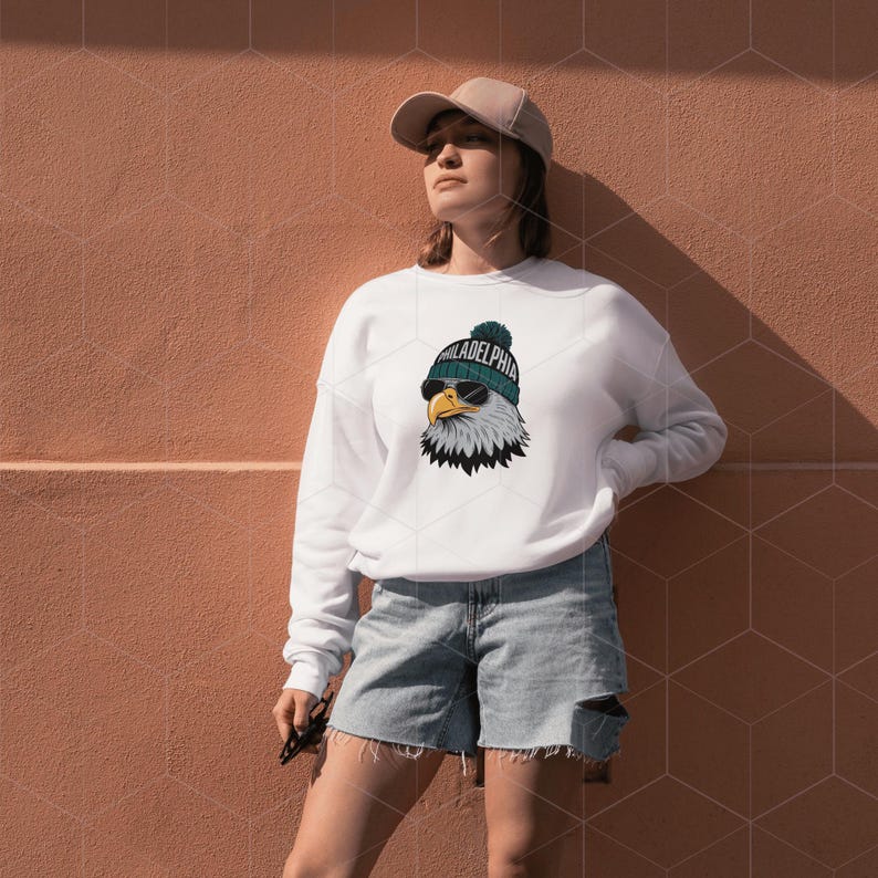 Peut inclure: Sweat-shirt blanc avec un graphique d'un aigle portant un bonnet vert et blanc avec le mot "Philadelphia" dessus. L'aigle porte des lunettes de soleil. Le mod&egrave;le porte un short en jean bleu et une casquette de baseball beige.