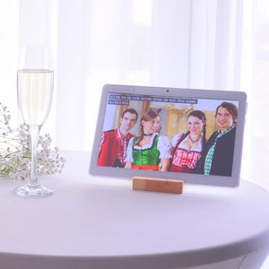 Può includere: Una donna brinda con un bicchiere di champagne mentre videochiama un gruppo di persone su un tablet. Il tablet è su un tavolo bianco con un mazzo di fiori bianchi.
