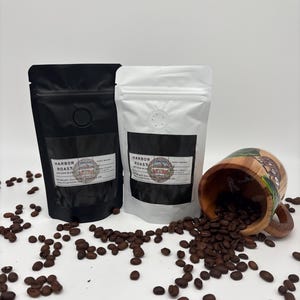 May include: Two sealed coffee bean bags, one black and one white, with labels reading "Harbor Roast Artisan Blend." A wooden mug, filled with coffee beans, is tipped over, spilling beans onto the surface. Scattered coffee beans complete the scene.