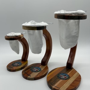 May include: Three wooden coffee filter holders with white cloth filters. The holders are made of different types of wood and have a curved handle. The base of each holder is oval shaped and has the words "Two Grounds" printed on it.